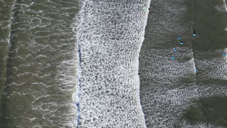 Aerial-view-of-foamy-waves-rolling-onto-a-dark-shore-with-scattered-surfers-waiting