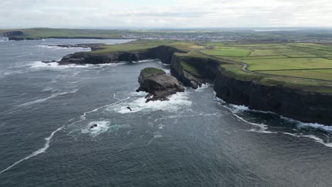 Kilkee-Cliffs-in-Ireland-with-waves-crashing-against-rocky-shores,-green-fields-in-the-distance,-aerial-view