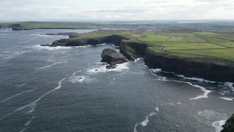 Irish-coastline-with-waves-crashing-against-cliffs,-green-fields-in-the-distance