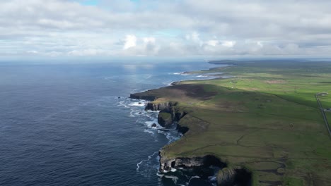 Rugged-coastline-with-waves-crashing-on-cliffs,-green-fields-alongside-the-ocean,-drone-footage