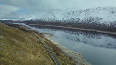 Aerial-tracking-shot-of-car-on-coastal-road-of-lake-in-scottish-highlands-with-snowy-mountaintop---Panorama-wide-shot