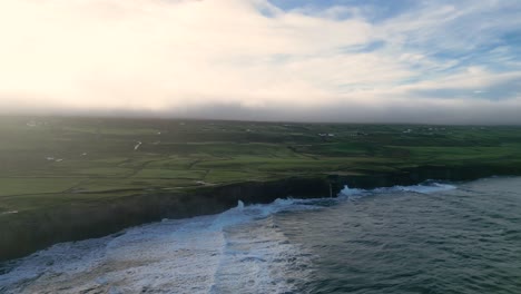 Rugged-coastline-with-lush-fields,-cliffs-and-ocean-waves-at-sunrise,-aerial-shot