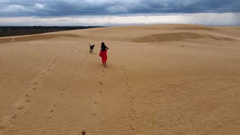 Aerial-follows-long-hair-Hispanic-woman-in-red-dress-on-sand-dune,-dog