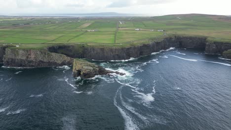 Dramatic-Kilkee-Cliffs-along-the-Irish-coast-with-lush-green-fields-and-ocean-waves,-aerial-view