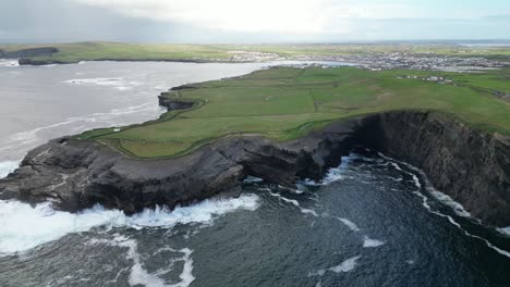 Dramatic-Kilkee-Cliffs-and-coastline-with-crashing-waves-and-green-fields,-under-a-cloudy-sky,-aerial-view
