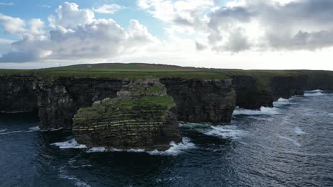 Kilkee-Cliffs-in-Ireland-with-waves-crashing-against-rocky-shores-under-a-cloudy-sky,-aerial-view