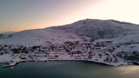 Snow-covered-village-by-the-sea-during-sunrise,-with-warm-light-on-the-horizon-at-ersfjordvegen
