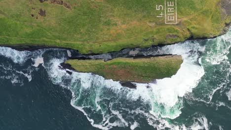 Rolling-aerial-view-of-rugged-coastline-with-'ÉIRE'-sign,-waves-crashing-against-cliffs