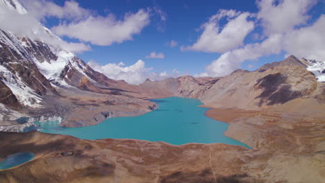 Drone-shot-of-World's-highest-lake-at-Annapurna-circuit-Nepal,-World's-highest-altitude-lake-Tilicho-surrounded-by-mountains-and-clouds,-blue-lake,-snow,-sunny-weather-adventure-4K