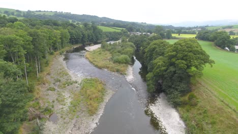 Drone-footage-flying-slowly-over-trees-and-a-slow-moving,-lazy-river-and-river-island-with-exposed-river-bed-and-vegetation-within-the-Lake-District,-UK