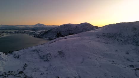 Verschneite-Landschaft-Bei-Sonnenuntergang-Mit-Bergen-Und-Ruhigem-Wasser,-Die-Warme-Himmelsfarben-Widerspiegeln