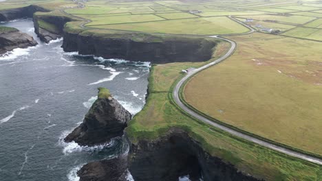 Aerial-view-of-Kilkee-Cliffs-in-Ireland,-winding-road-along-rugged-coastline,-green-fields-on-a-cloudy-day