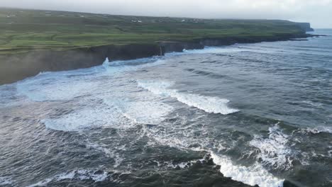Doolin's-rugged-coastline-with-waves-crashing-against-cliffs,-green-fields-in-the-distance,-aerial-view
