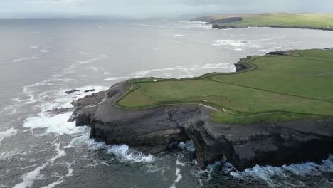 Kilkee-coastline-with-grassy-cliffs-and-waves-crashing-against-the-shore,-aerial-view