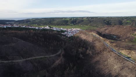 Drone-flyover-burnt-forest-area-by-small-village-in-Algarve,-flying-backward