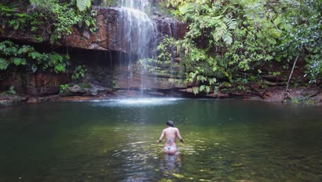 Woman-in-tiny-bikini-enters-calm-pool-below-pretty-jungle-waterfall