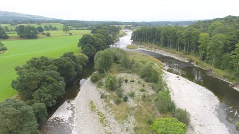 Beautiful-drone-footage-flying-over-a-tree-lined-shallow,-slow-moving-river-and-river-island-in-the-rural-countryside-of-the-Lake-District,-UK