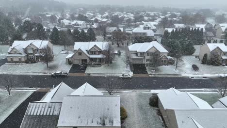 A-suburban-street-with-two-story-houses-under-a-light-snowfall,-roads-partially-covered,-and-bare-trees