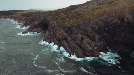 Aerial-view-from-drone-flying-toward-rocky-shoreline-with-waves-breaking-against-cliffs
