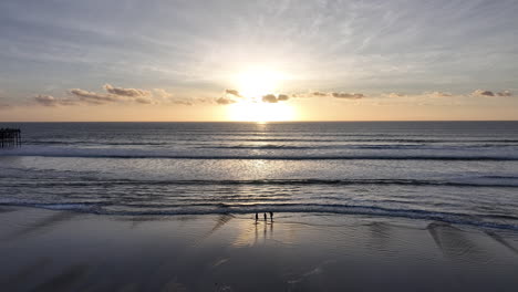 Drone-shot-orbiting-around-two-people-walking-at-ocean-shoreline-at-sunset
