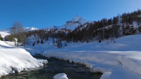 Drone-flying-at-low-altitude-over-mountain-stream-flowing-through-snowy-landscape-on-sunny-day,-Italian-Alps