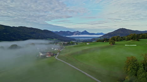 Light-fog-cloud-over-village-in-green-fields-of-alpine-mountain-valley
