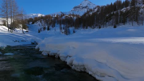 Drone-flying-low-over-stream-flowing-through-snow-covered-and-frozen-Valmalenco-on-sunny-day,-Alps-in-Italy