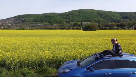 revealing-shot-man-chilling-sitting-on-roof-of-a-blue-car-in-bright-colorful-yellow-green-field-of-flowers-relax