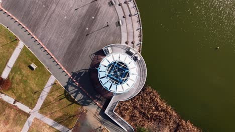 A-top-down-shot-over-a-circular-building-next-to-a-lake-on-a-sunny-day-in-a-park