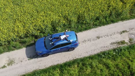 internal-freedom-of-a-man-chilling-sitting-on-roof-of-a-blue-car-in-yellow-green-field-of-flowers---zoom-out-drone-shot