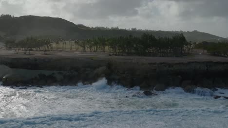 Drone-dolly-above-ocean-waves-crashing-on-rocky-shoreline-with-coconut-trees-on-cliff-edge