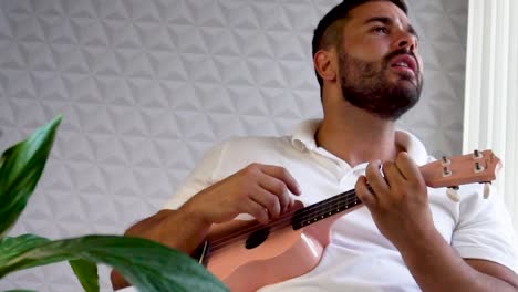 Man-mid-30-and-with-beard-is-playing-and-singing-with-his-pink-ukulele-next-to-a-big-window-and-close-to-a-green-plant
