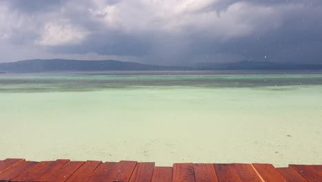 Rain-downpour-onto-wooden-wharf-in-the-tropics-overlooking-turquoise-ocean-water-on-tropical-island-in-Raja-Ampat,-West-Papua,-Indonesia