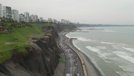 Drone-widens-the-lens-over-Miraflores,-Lima:-Coastal-highway,-housing-towers-atop-the-cliff