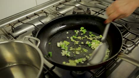 Close-up-of-chopped-green-onions-being-sautéed-in-a-frying-pan-on-a-gas-stove-by-a-person-with-a-spatula