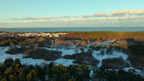 Aufsteigende-Drohnenaufnahme-Einer-Waldlandschaft-Mit-Schnee-Und-Danziger-Stadtteil-Im-Hintergrund-Bei-Sonnenaufgang---Blaue-Ostsee-Im-Hintergrund