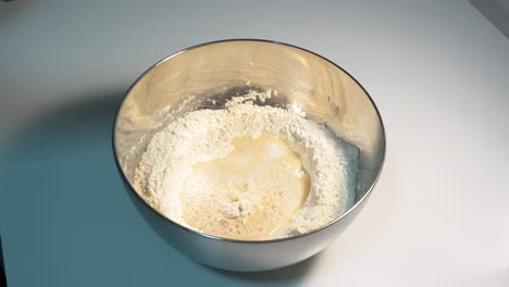 Mixing-Ingredients-in-a-Stainless-Steel-Bowl-on-a-White-Tabletop,-Pizza-Dough,-Bread-dough