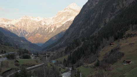 Alpine-valley-with-snow-capped-mountains,-a-train-curving-along-tracks,-highway,-and-scattered-houses-at-dusk
