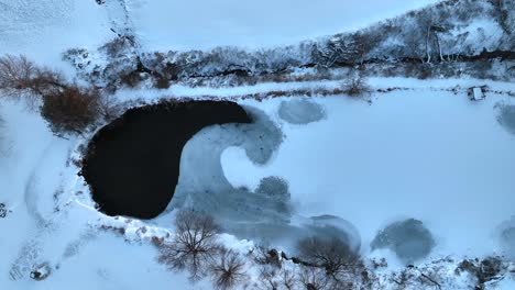 Top-down-aerial-of-farm-pond-partially-frozen-and-covered-in-winter-snow-and-ice
