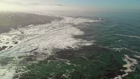High-flying-aerial-view-of-ocean-and-land-as-waves-roll-in-while-panning-up-from-waters-to-display-shoreline-with-a-bit-of-mist