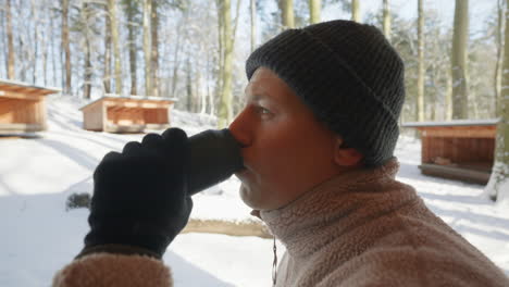 Close-up-of-a-person-in-winter-attire-drinking-from-a-thermos-outside-snowy-cabins