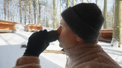 Profile-view-of-a-person-sipping-from-a-thermos-cup,-with-snowy-woodland-cabins-in-the-background