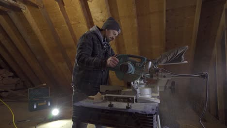 Man-in-beanie-using-a-miter-saw-in-a-woodshop,-sawdust,-focused-craftsmanship,-indoor-lighting