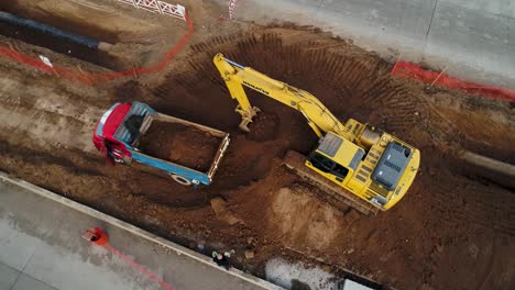 Construction-site-with-excavator-loading-dirt-into-dump-truck,-busy-workday,-aerial-view
