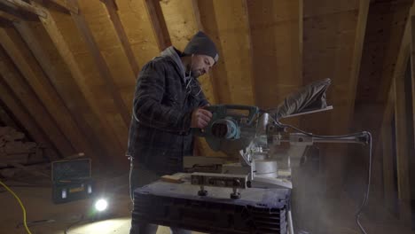 Man-in-beanie-and-jacket-using-a-mitre-saw-in-a-wooden-workshop,-dust-in-the-air,-indoor-lighting