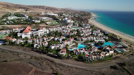 Coastal-resort-in-Fuerteventura-with-white-buildings,-pools,-next-to-a-sandy-beach,-daytime,-aerial-view