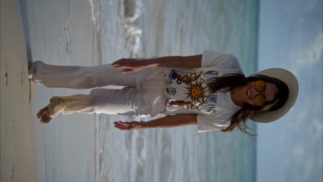 Vertikale-Zeitlupe-Einer-Schönen-Lateinamerikanischen-Brünetten-Frau,-Die-Am-Strand-In-Richtung-Kamera-Läuft-Und-Leinenhosen,-Einen-Weißen-Hut-Und-Eine-Gelbe-Sonnenbrille-Trägt