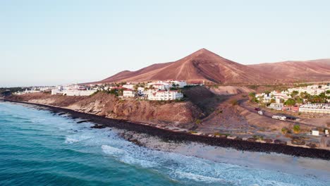 Fuerteventura-coastline-with-volcanic-mountain,-ocean-waves,-and-coastal-town-at-sunrise,-aerial-view