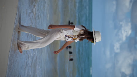 Vertical-slow-motion-of-a-beautiful-latin-brunette-woman-walking-on-the-beach-wearing-linen-pants-a-white-hat-and-yellow-sunglasses-enjoying-the-sea