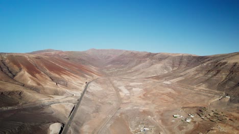 Desert-road-winding-through-the-barren,-hilly-landscape-of-Fuerteventura-under-a-clear-blue-sky,-aerial-view